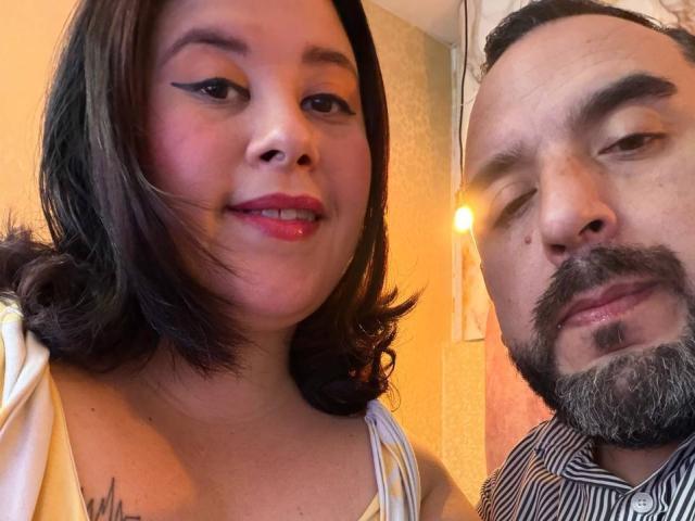 A SarahMat tem cabelos pretos e brilhantes e batom vermelho, vestindo uma blusa amarela. Ela está ao lado de um homem de barba com uma camisa listrada, e ambos estão sorrindo para a câmera..