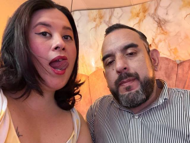 SarahMat, avec ses courts cheveux bruns, son rouge à lèvres rouge, la langue tirée, montrant un petit tatouage sur sa poitrine, à côté d'un homme barbu en chemise rayée, qui a l'air légèrement agacé..