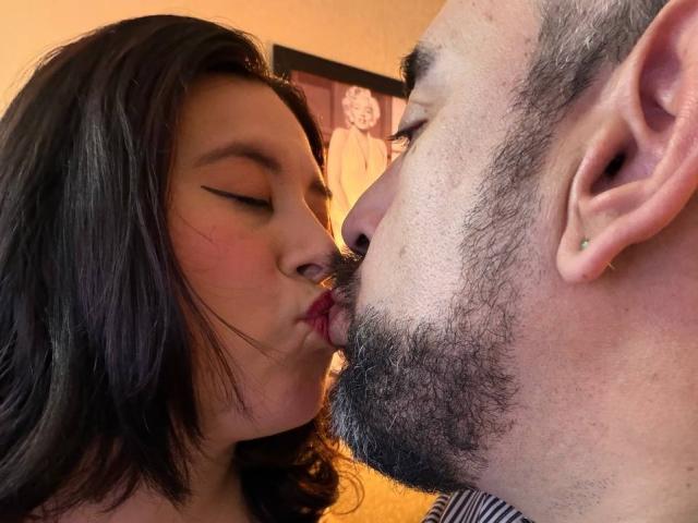 SarahMat and a bearded dude are kissing with their eyes closed. SarahMat has dark hair and winged eyeliner, while the dude has a beard and ear piercing.