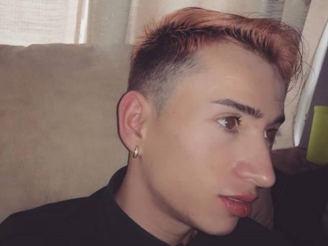 IamSmiith a les cheveux courts, teints en orange, une coupe sur le côté et une boucle d'oreille dorée. Il/Elle porte un haut noir, regarde vers la droite avec une expression sérieuse..
