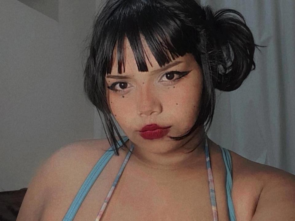 Majudy has black hair in a messy bun, bold eyeliner, red lipstick, freckles, and a blue halter top, showing a serious expression, with a slight pout on her lips.