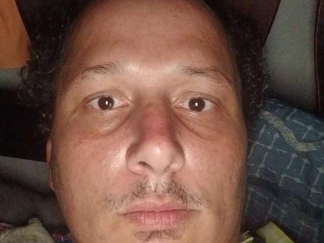 Close-up of Macknd6.9's face, centered in the frame. His medium brown skin is slightly flushed, with visible pores and a subtle sheen. Macknd6.9 has a balding forehead with curly, dark brown hair on the sides. He has small, brown eyes, a rounded nose, and a thin mustache with sparse hair around his lips. The background is blurry, with a blue patterned fabric on the right and an orange object on the left. The lighting is soft, highlighting his facial features.