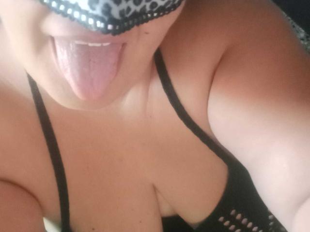 PassionD with her tongue out, wearing a leopard-print mask and black mesh bra, showing her big, round boobs and tan skin. Her tongue is pink and slightly wet.