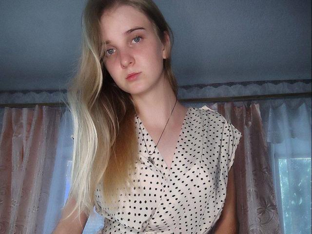 SiliensXRouses with blonde hair, blue eyes, and a polka dot dress, has small tits, a necklace, and a serious expression in a well-lit room with pink curtains.