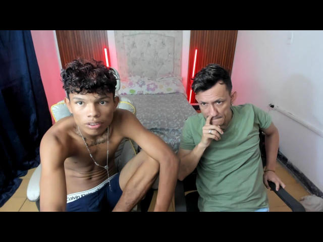 Two men in a room, JuanAndLuke on the left is shirtless, wearing black boxer shorts, with curly brown hair and a necklace. He leans forward, looking at the camera. Luke on the right, in a green t-shirt, sits with a finger to his lips, looking at JuanAndLuke. Red vertical lights are in the background, and a bed with floral patterns is behind them. The room has white walls and a wooden floor. The image has a casual, realistic style.