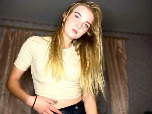 SiliensXRouses with blonde hair, fair skin, and blue eyes, wearing a beige ribbed crop top, black pants, and a black bracelet, looking slightly to the side.