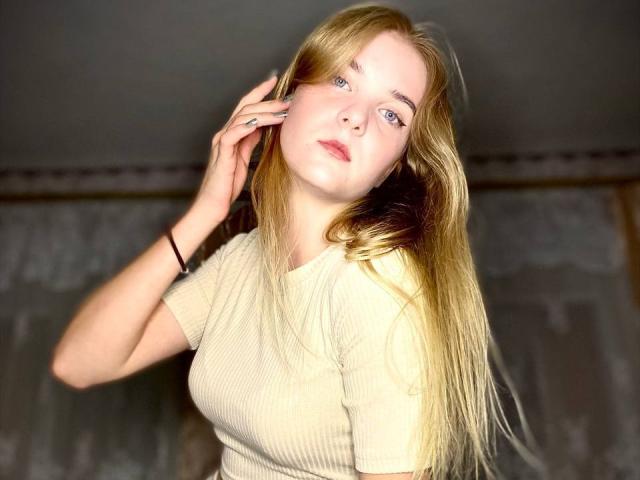 SiliensXRouses has long blonde hair, blue eyes, and is wearing a beige ribbed shirt, with one hand touching her head. Her tits are small and she looks slightly bored.