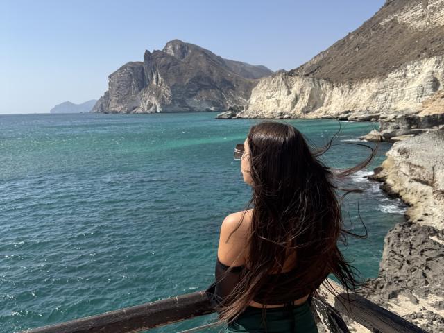 A photograph of a woman, BlueAriana, with long dark brown hair blowing in the wind, wearing sunglasses and a black strapless top. She stands on a wooden railing overlooking a turquoise sea with rocky cliffs and a large, rugged mountain in the background. The sea has gentle waves, and the clear sky is blue. The rocky coastline on the right has a textured, uneven surface. BlueAriana's back is to the camera.