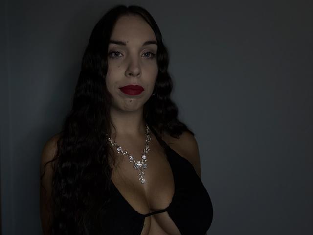 BlueAriana with long dark wavy hair, wearing a black halter top showing her large tits, and a sparkling necklace, has red lipstick and a slight bruise on her cheek.