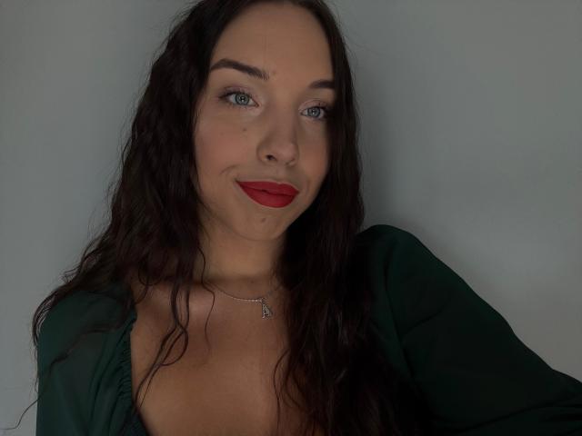 BlueAriana has wavy dark brown hair, red lipstick, and a green top, showing her tits in the photo. She's wearing a necklace with an "A" pendant.