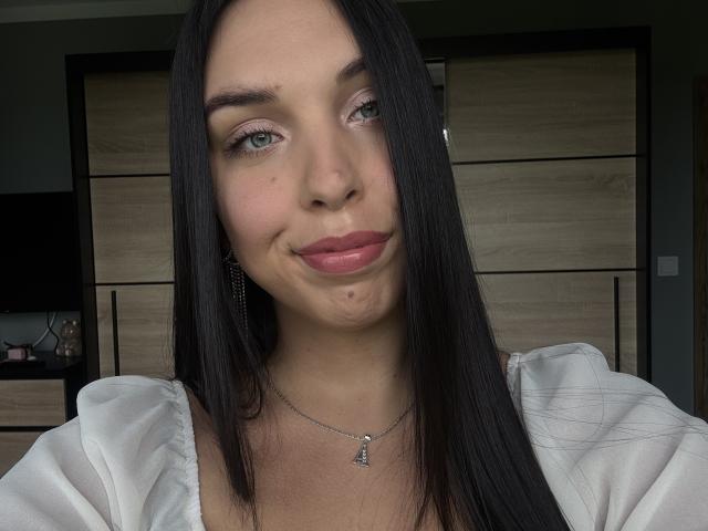 Close-up of BlueAriana with long straight black hair, green eyes, and pinkish lipstick. She's smiling slightly with a small dimple on her right cheek. She wears a white shirt with puffed sleeves and a silver necklace with a small pendant. Her skin is light with a few small blemishes. The background is a wooden cabinet with black handles. The lighting is natural, highlighting her facial features and hair. The image has a casual, realistic style.