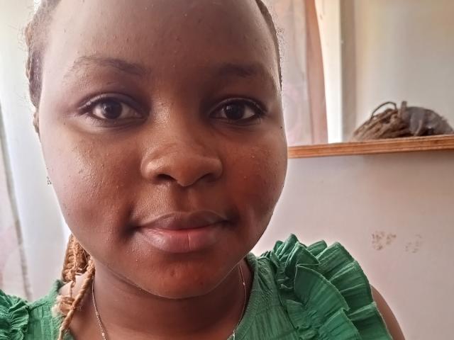 Pinacol's face, brown skin, small nose, dark eyes, green ruffled dress, slight smile, braided hair, light brown wall in background, small brown basket on wooden shelf to the right.
