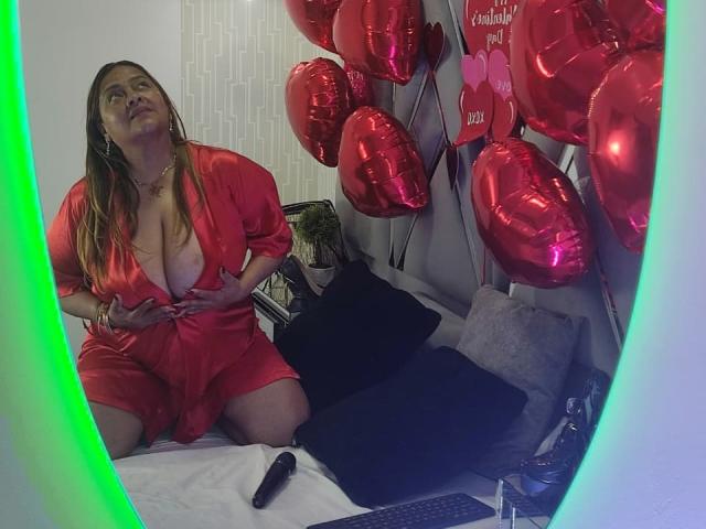 MonzerratBoyer kneels on a bed wearing a red, low-cut dress, revealing her big, bare tits. Red, shiny balloons surround her, with a black vibrator on the bed.