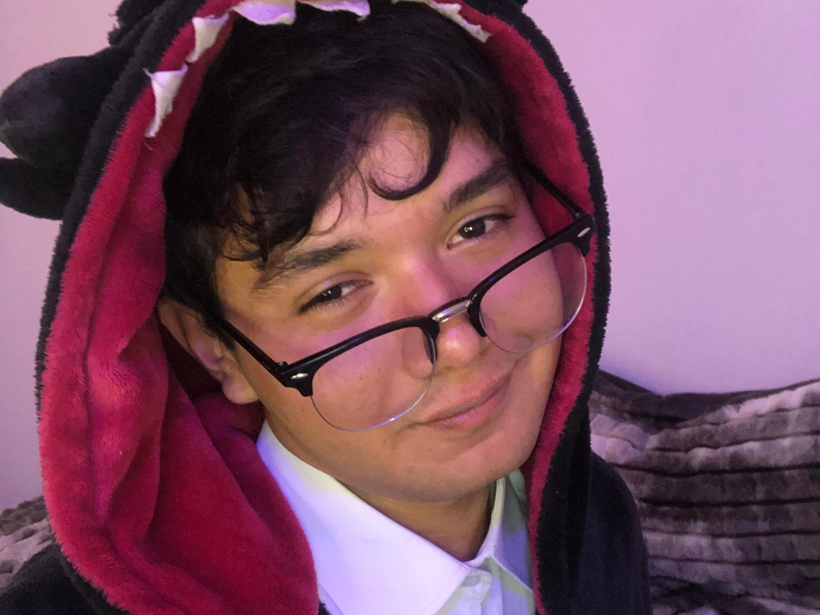 PrinceKody, a cute dude with dark messy hair and glasses, wears a black and red fluffy dragon hood, smiling softly at the camera, looking slightly nerdy and adorable.