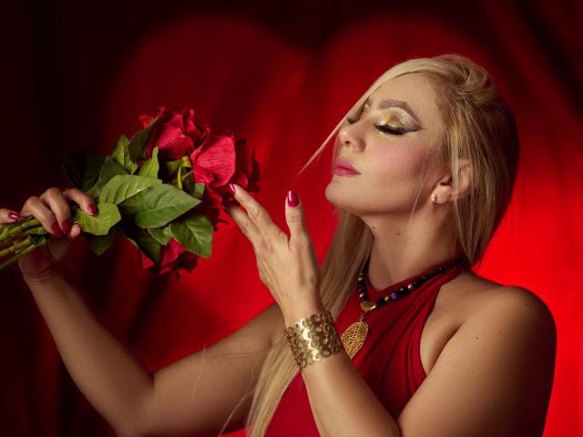 RoxannEmilss with blonde hair, red dress, gold bracelet, and purple nail polish, smelling red roses, eyes closed, red background, wearing a necklace with a red gem.