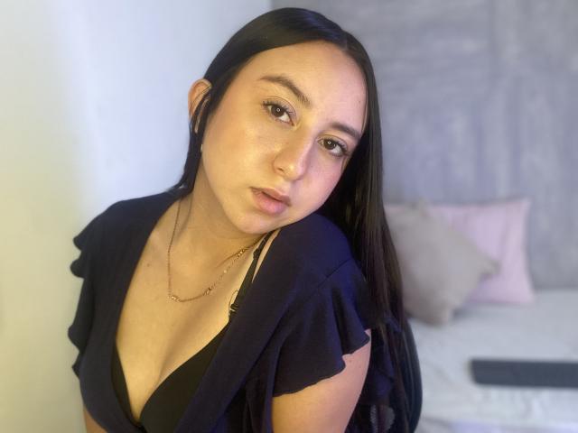 RosalieDupont with long black hair, wearing a black top, showing her tits in a low-cut, has a gold necklace, and a slightly confused expression, in a bedroom.