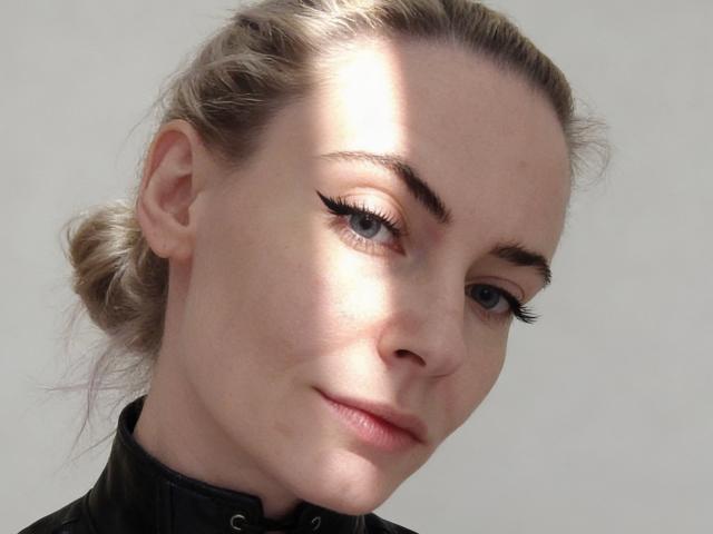 SharonJackkson a la peau claire, les yeux bleus, et un trait d'eye-liner noir avec des cils épais. Elle porte une veste en cuir noir, et ses cheveux blonds sont attachés en chignon. Elle regarde vers le bas, avec un léger sourire..