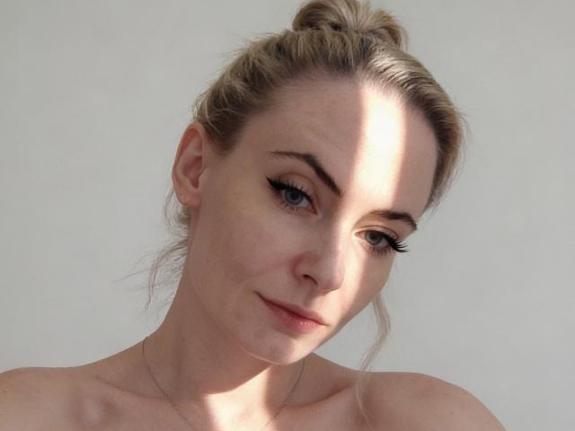 SharonJackkson hat helle Haut, blondes Haar zu einem unordentlichen Dutt gebunden, und schwarzen Eyeliner mit langen Wimpern. Sie schaut zur Seite und hat ein leichtes Lächeln. Sie ist oben ohne..