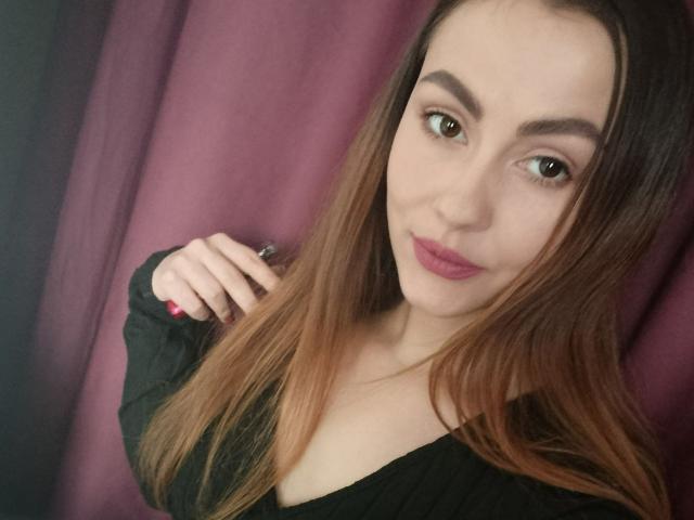 KendyOwers69 with long brown hair, brown eyes, pink lipstick, and black top, holding hair near her ear, showing her tits and a slight smile.