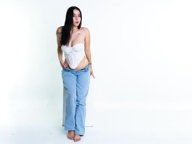 SammyRodriguez with long black hair, wearing a white strapless corset and loose blue jeans, standing barefoot, showcasing her tits and ass, with a small tattoo on her chest.