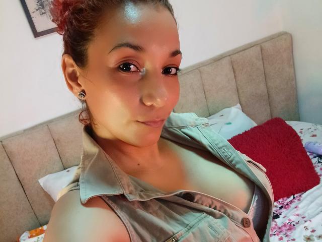 AmberDemons, a curvy woman with tan skin, small black earrings, and a denim sleeveless vest, showing her tits, red curly hair, and a slight smile in a bedroom.