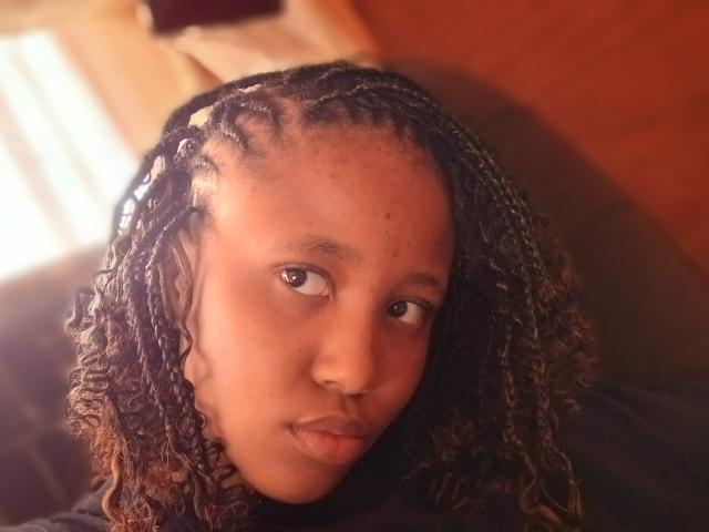 Close-up of CuteLarona, a young woman with dark brown skin and curly, braided hair. Her hair is styled with small white beads at the roots. She has a neutral, slightly pouty expression with her lips slightly pursed. Her eyes are dark and focused on the camera. The lighting is warm, casting a soft glow on her face. She is wearing a dark-colored shirt. The background is blurred, with a brown wall and a window with white curtains. The image has a natural, candid feel.