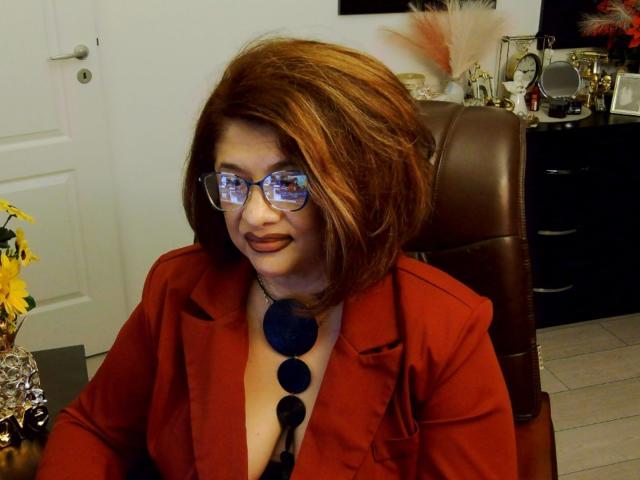 Elegante pero atrevida, con cabello rojo, gafas y un blazer rojo, mostrando el escote con un collar azul, sentada en una silla de cuero marrón, sonriendo con lápiz labial marrón..