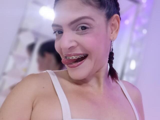 KatteRouse has metal teeth and is smiling with her tongue out, wearing a white tank top, and has dark braided hair, showing a cute and slightly edgy look.