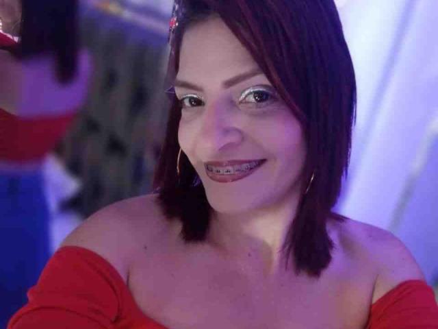 KatteRouse with red off-shoulder top, smiling, showing metal teeth, wearing hoop earrings, dark red bob, slight blue-purple light. Boobs partially visible, face centered.