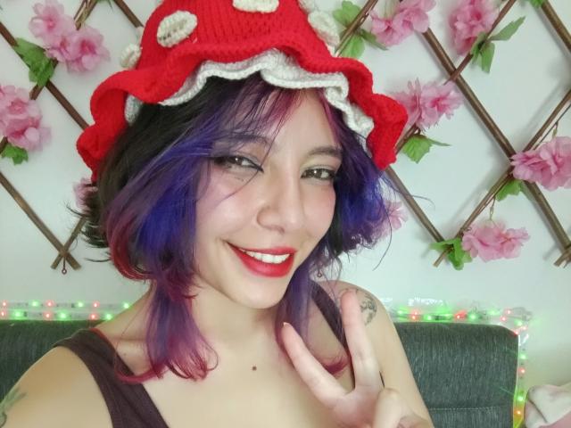 DarckVelvet with purple and black hair, wearing a red mushroom hat, smiling with red lipstick, black tank top, showing a finger with a tattoo, and purple highlights.