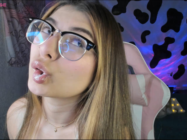 AbbyDew with glossy lips, puffy pink lipstick, and clear glasses, pouting with a small tongue out, wearing a gold necklace, sitting in a pink chair.
