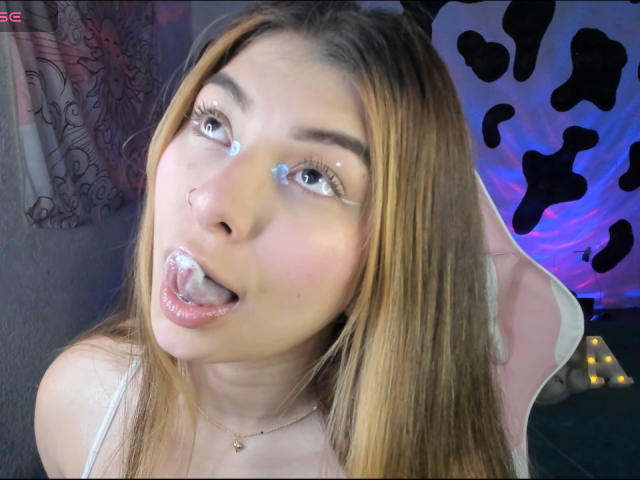AbbyDew has cum on her face and tongue, with white streaks in her brown hair, looking up with her mouth open. She's wearing a white top and a necklace.