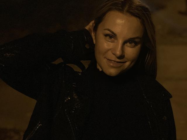 BelleLisaG has a sly smile, brown eyes, and light skin, wearing a black lace jacket, with her hand in her hair, in dim, warm lighting.