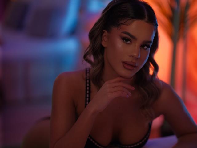 NikkiPearl, a young woman with wavy brown hair, sits in a dimly lit, colorful room. She has a light skin tone, full lips, and dark, defined eyebrows. She wears a black, strappy bra that reveals her cleavage. Her right hand rests gently on her chin. The light casts a blue and orange glow on her, highlighting her smooth, slightly shiny skin. The background is blurred, emphasizing NikkiPearl's relaxed, confident expression. Her makeup is subtle yet effective, with a focus on her eyes.