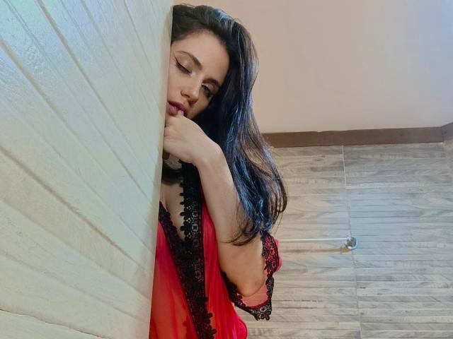 DanaLamiri with long dark hair, red and black lace dress, leaning against a white brick wall, her hand near her mouth, looking down seductively, showing her tits.