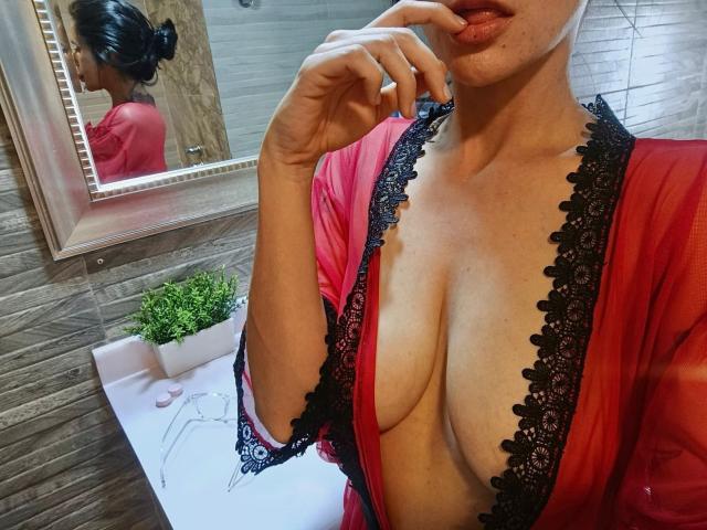 DanaLamiri in a red, lace-trimmed, sheer robe, exposing her large, bare tits. She's biting her finger, with a reflection in the mirror showing her black hair bun.