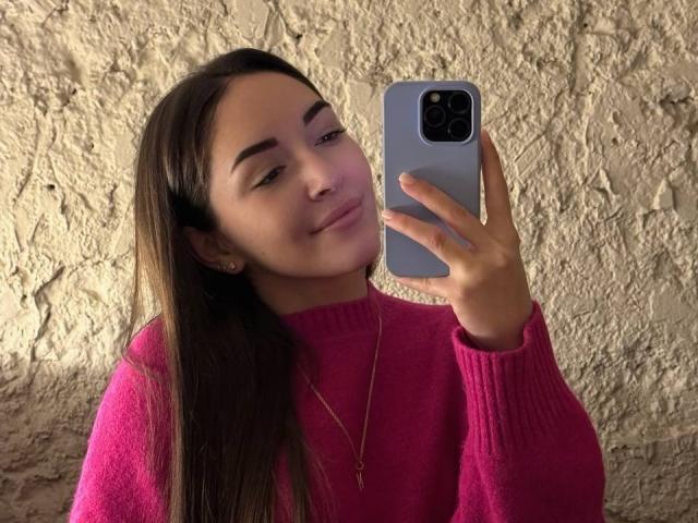 AquaLady with long brown hair, pink sweater, and gold necklace, taking a selfie with a blue iPhone, smiling slightly, against a textured beige wall.