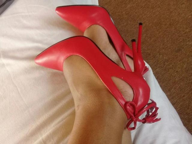 VanessaMoabi-hot's feet in red, pointy, high-heeled shoes with cut-out sides and a ribbon bow, resting on a white bed. Her skin is smooth and slightly tanned.