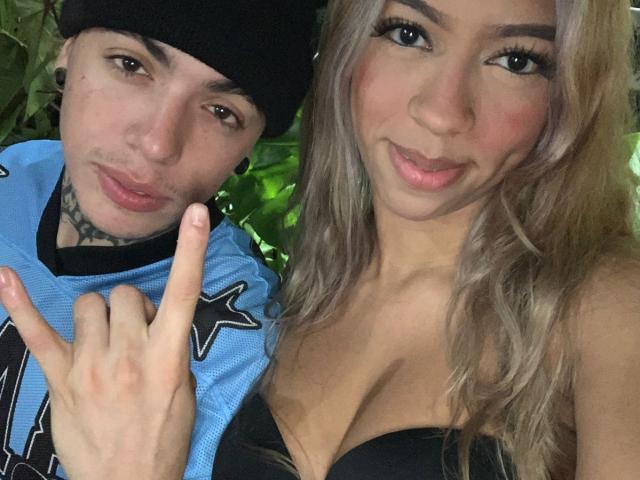 Close-up selfie of IceCumHot. On the left, a man with a black beanie, brown eyes, and a tattoo under his chin wears a blue sports jersey with black star details. He holds up his middle finger. On the right, a woman with long, wavy blonde hair, pink lips, and dark eyebrows wears a black strapless top, showing cleavage. Her right arm is around the man. The woman's expression is slightly smiling. The man's expression is neutral. The lighting is even.