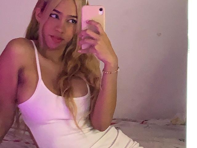 IceCumHot with blonde hair, wearing a white tank top, showing her tits, holding a pink phone, smiling, pink lighting, sitting on a bed.