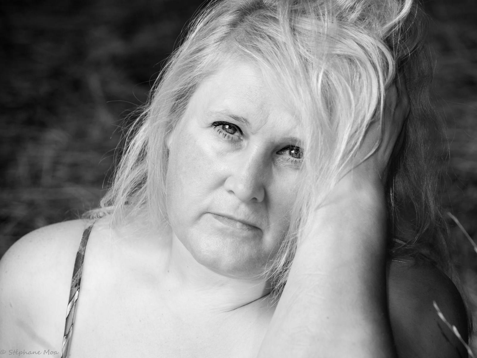 Black-and-white photo of CarpediemLife with messy blonde hair, fair skin, and small tits, looking slightly sad, hand in hair, wearing a thin-strapped top, watermark in bottom left.