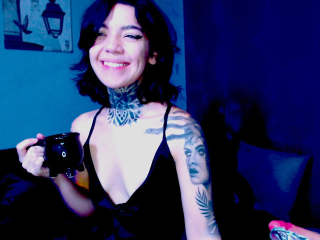 LannieGonzales with black hair, tattoos on neck and arm, holding a black cup, wearing a low-cut black top, smiling, blue-lit room, showing tits, short bob hairstyle.