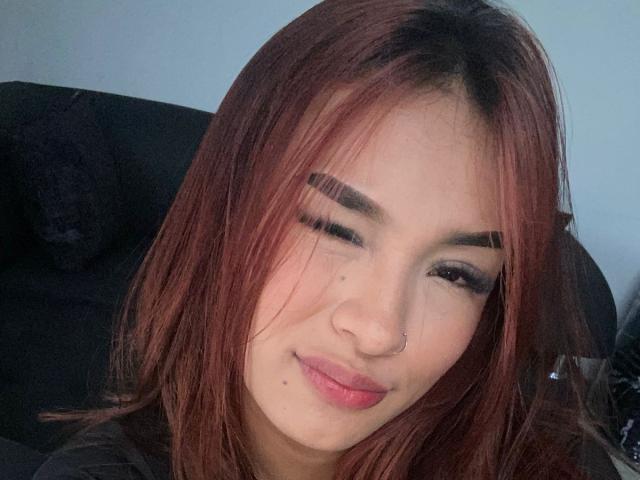 SashaHans has reddish-brown hair, thick eyebrows, dark eyeliner, a nose ring, and a slight smile with a small mole above her upper lip. She's winking.