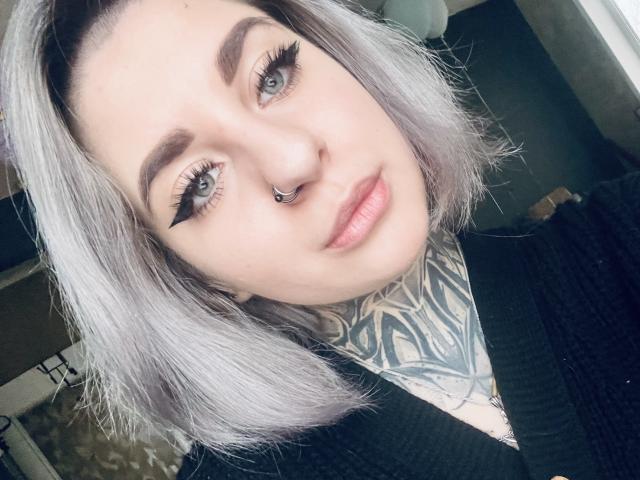 AllisonXAlfred with silver hair, nose ring, black eyeliner, tattooed neck, and pink lips, wearing a black top. Her tits are not visible. Her ass is not visible.