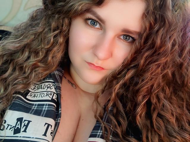 MinistraX with curly brown hair, blue eyes, and pale skin, wearing a plaid shirt, showing her large, round tits, and a small tattoo on her chest.