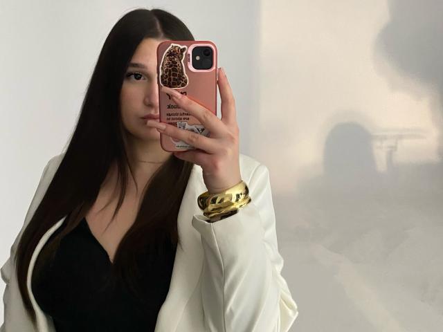 MissKuromi takes a mirror selfie. She has long, straight brown hair and a fair complexion. She holds a pink phone case with a leopard print design in her right hand. She wears a white jacket over a black V-neck top and a gold bracelet on her left wrist. The phone's case has small text and a sticker on the back. Her shadow is visible on the right. The image has a soft, diffused light. She looks directly into the camera with a neutral expression.