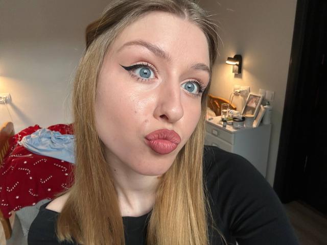 Nikollia has blue eyes, black eyeliner, pink lipstick, and blonde hair. She's making a pouty face in a black top. Her ass is not visible.