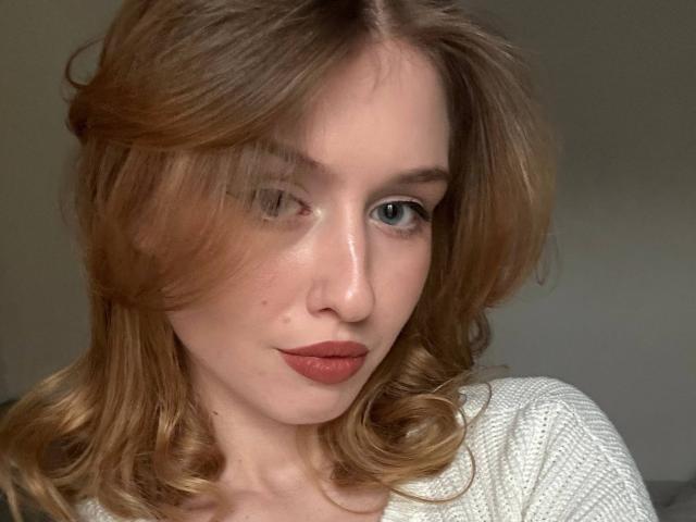 Nikollia with wavy, reddish-brown hair, blue eyes, and red lipstick, wearing a white knitted top, showing a subtle smile and a small mole on her left cheek.
