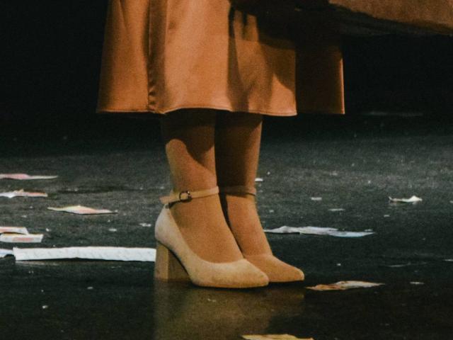 Nikollia's feet in tan, suede, high-heeled shoes with a small strap and block heel are centered. Her orange, knee-length skirt partially covers her legs. The dark, textured floor is scattered with small, colorful paper pieces. The light focuses on her shoes, highlighting their texture and color. The composition emphasizes the contrast between the shoes and skirt.
