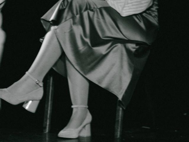 Nikollia's legs crossed, wearing a shiny satin skirt and ankle-strap high heels with chunky heels, sitting on a chair. Her anklet is visible, and the image is black and white.